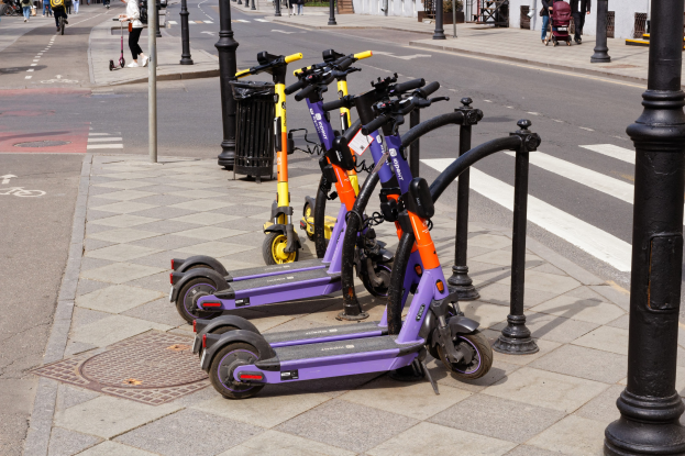 Eine Reihe von Elektroscootern, die entlang einer Straße mit Fußgängern, Radfahrern, einem Müllcontainer im Vordergrund und Gebäuden, Bäumen und Pflanzen im Hintergrund geparkt sind.