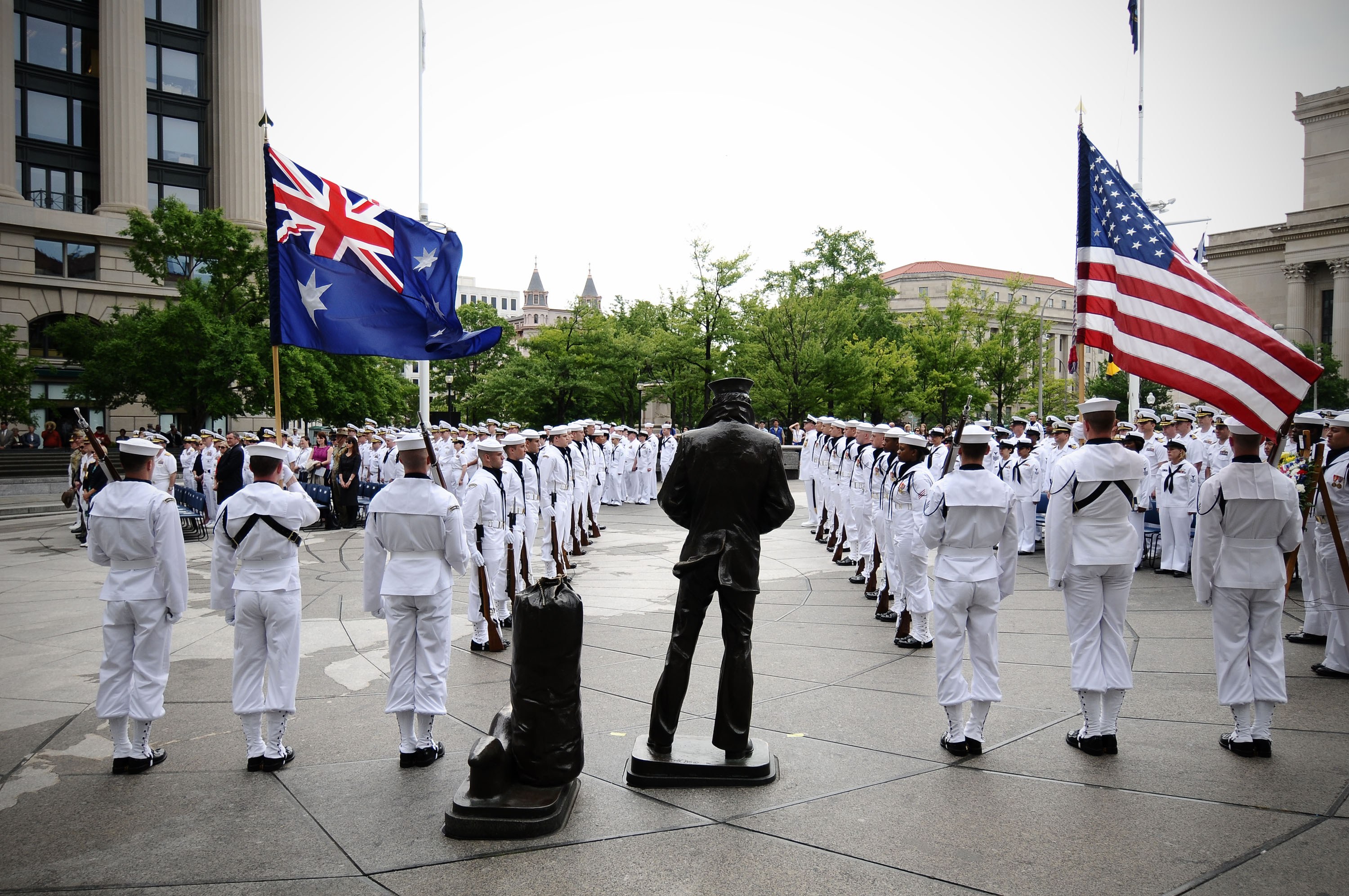 Eine Gruppe von Männern in weißen Uniformen mit Mützen und Gewehren steht in Formation vor Flaggen, mit einer Statue im Vordergrund, Bäumen und Gebäuden im Hintergrund und Menschen, die auf Stufen unter einem klaren blauen Himmel sitzen.