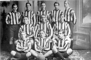 Ein Schwarz-Weiß-Foto einer Fußballmannschaft, bei der einige Spieler stehen und andere auf dem Boden sitzen, ein Pokal in der Mitte, Säulen und eine Wand im Hintergrund und Text oben und unten auf dem Foto.