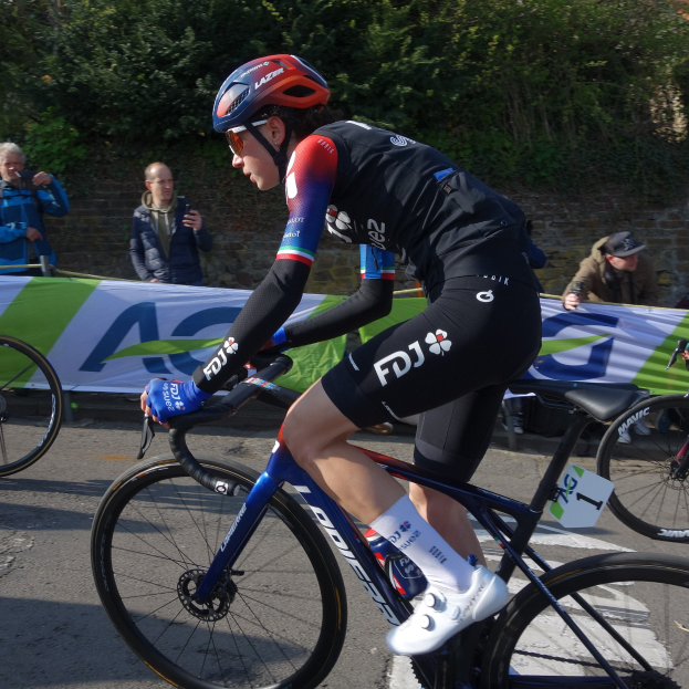 Die professionelle Radsportlerin Katie Archibald fährt mit Helm, Schutzbrille, Handschuhen und Schuhen auf einem Fahrrad über eine Straße neben einer Menge von Menschen. Im Hintergrund gibt es ein Banner mit Text, eine Wand, Bäume und ein Gebäude.