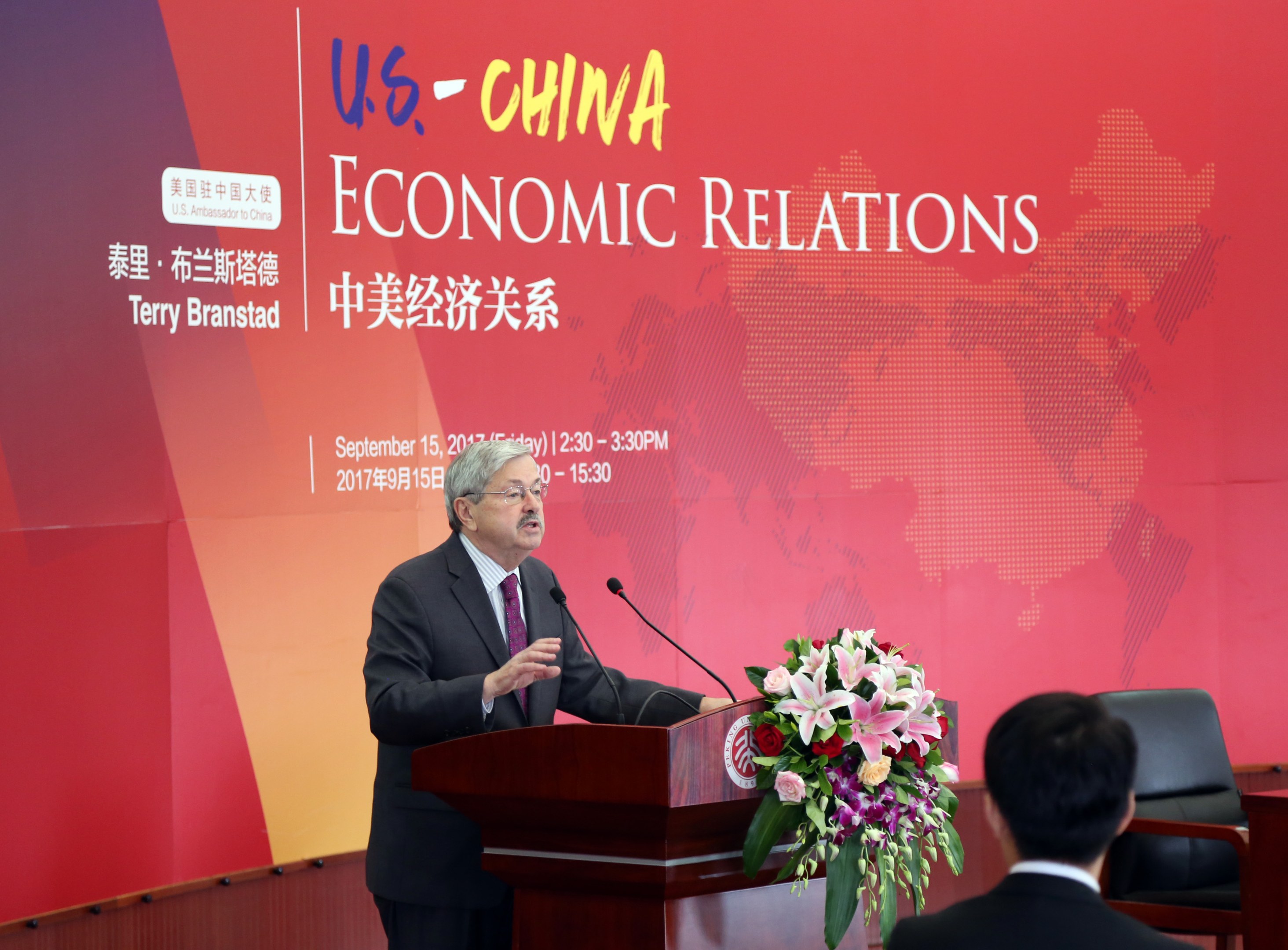 Ein Mann in einem schwarzen Blazer, weißem Hemd und lila Krawatte steht an einem Podium mit zwei Mikrofonen und einem Blumenstrauß, während er vor einer roten Wand mit einem roten Banner spricht, auf dem "US-China Economic Relations" steht. Rechts von ihm sitzt eine weitere Person auf einem Stuhl.