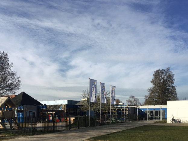 Großes Gebäude mit Metallzaun, Rasenfläche, Weg, Bäumen, Schildern, einem abgestellten Fahrrad, nächsten Häusern und einem bewölktem Himmel, identifiziert als neuer Kinderspielplatz an der Schule.