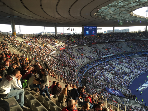 Große Zuschauermenge im Allianz Arena Stadion bei einem Fuballspiel mit einer Bühne, Fahnen und einem Bildschirm im Hintergrund.