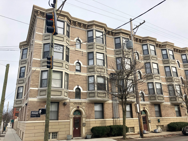 Großes Wohnhaus an einer Straßenecke mit der Aufschrift "Park Apartments for Rent in Chicago, IL"-Schild, Bäumen, Topfpflanzen, Strommasten, einer Ampel, einem Auto und einem bewölkten Himmel.