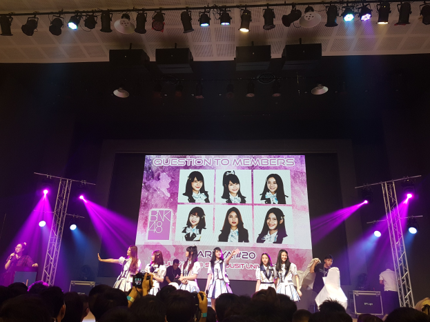 Eine Gruppe von Menschen, die auf einer hell erleuchteten Bühne spielt, mit einer Menge darunter, mit einem Menschen mit Mikrofon und einem anderen, der ein Instrument spielt, und einem großen Bildschirm mit BK48 im Hintergrund.