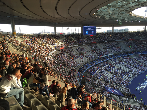 Eine große Menschenmenge sitzt im Allianz Arena-Stadion in München, Deutschland, und schaut ein Fußballspiel. Rechts im Bild befindet sich eine Bühne mit ein paar Menschen darauf, und im Hintergrund sind Fahnen, Stangen und ein Bildschirm zu sehen. Der Himmel ist oben im Bild zu sehen, und das Stadion scheint das Allianz Arena in München, Deutschland, zu sein.
