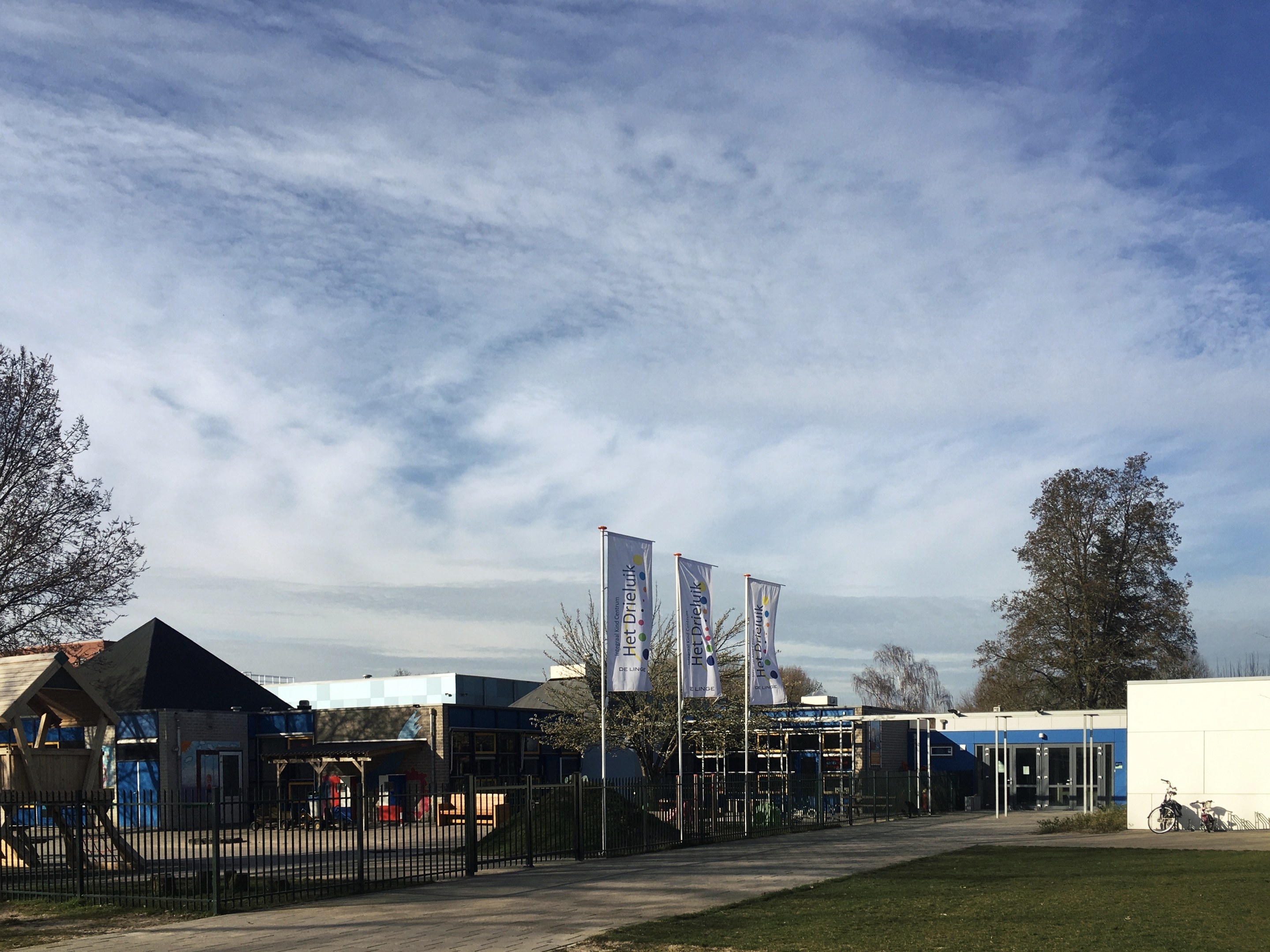 Großes Gebäude mit einem großen Rasen vorne, umgeben von einem Metallzaun, Bäumen, Bannern, einem Fahrrad, nahen Häusern und einem bewölkten Himmel, als neuer Spielplatz an der Schule identifiziert.