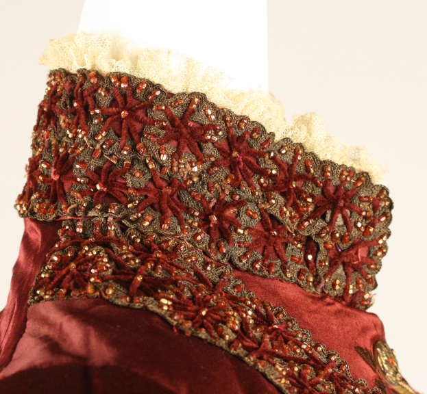 Nahaufnahme eines roten und goldenen Kleides mit aufwändiger Spitzenverzierung, das auf einem Mannequin vor einem weißen Hintergrund präsentiert wird.