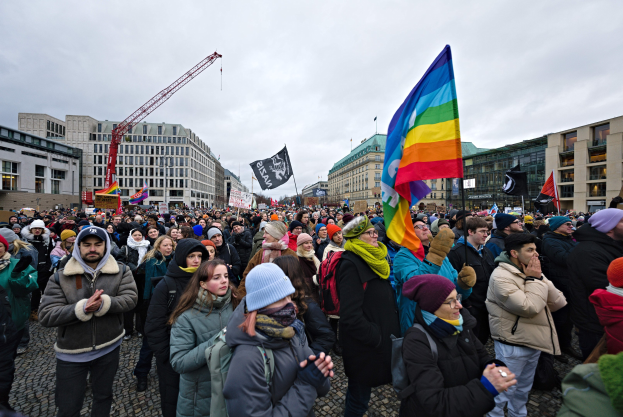Eine große Gruppe von Menschen mit Fahnen und Schildern steht vor einem Gebäude während einer LGBTQ+-Rechtsdemo in Berlin, mit Gebäuden, einem Kran und Wolken im Hintergrund.