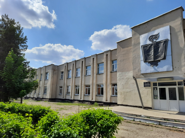 Großes Gebäude mit einer Tafel an der Seite, umgeben von Bäumen und einer Straße, unter einem bewölkten Himmel, identifiziert als Tschernobyl-Kernkraftwerk.