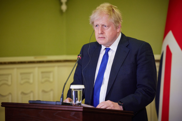 Ein Mann in Anzug und Krawatte spricht an einem Podium mit Mikrofon, Glas Wasser und anderen Gegenständen, vor einer Wand mit einer Flagge auf der rechten Seite.