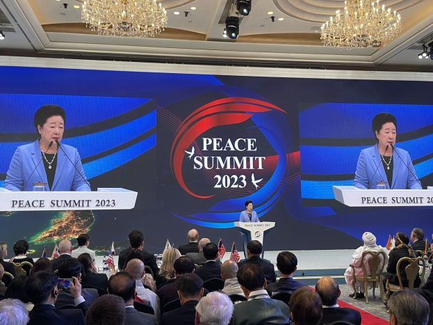 Zwei Frauen stehen auf Podien mit Mikrofonen und sprechen zu einem sitzenden Publikum auf dem Peace Summit in Hong Kong, wobei ein Bildschirm mit Text und Logos im Hintergrund zu sehen ist.