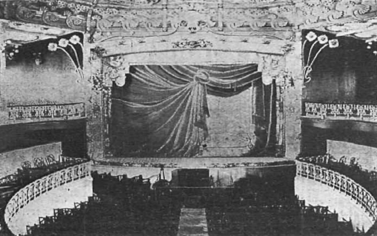 Schwarzes und weißes Foto des Theatersaals des Theatre Royal in London, das eine Bühne mit einem Vorhang, Geländern und verzierten Wanddesigns zeigt, mit ein paar sitzenden Besuchern und Text am unteren Rand.
