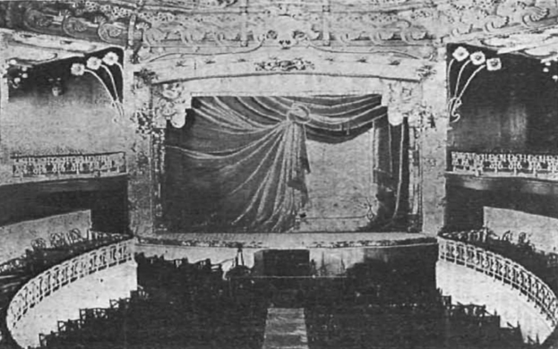 Schwarzes und weißes Foto des Theatersaals des Theatre Royal in London, das eine Bühne mit einem Vorhang, Geländern und verzierten Wanddesigns zeigt, mit ein paar sitzenden Besuchern und Text am unteren Rand.
