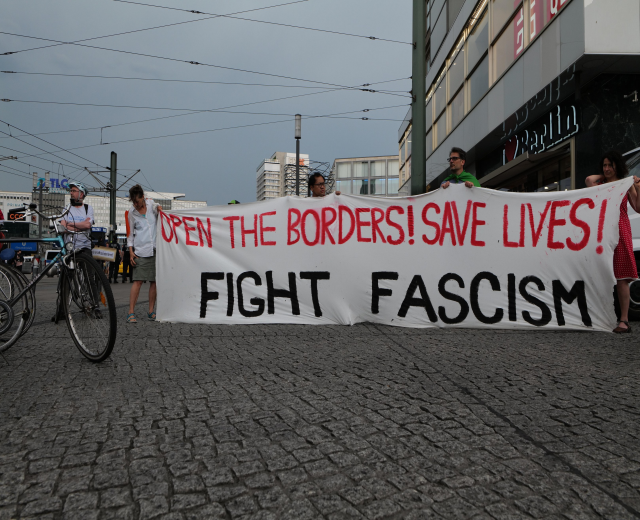 Gruppe von Menschen mit einem Banner 'Öffnet die Grenzen, rettet Leben, kämpft gegen Faschismus' auf einer Straße mit Fahrrädern auf der linken Seite, Gebäuden und Strommasten im Hintergrund unter einem klaren blauen Himmel.