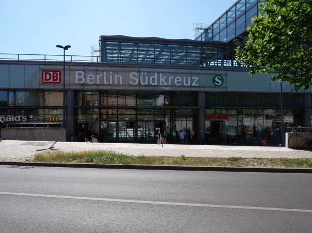 Großes gläsernes Berliner Südkreuz-Bahnhofgebäude mit Text an seiner Fassade, umgeben von Straßeninfrastruktur, Fahrzeugen, Fußgängern, Fahrrädern, Bäumen und einem klaren Himmel.