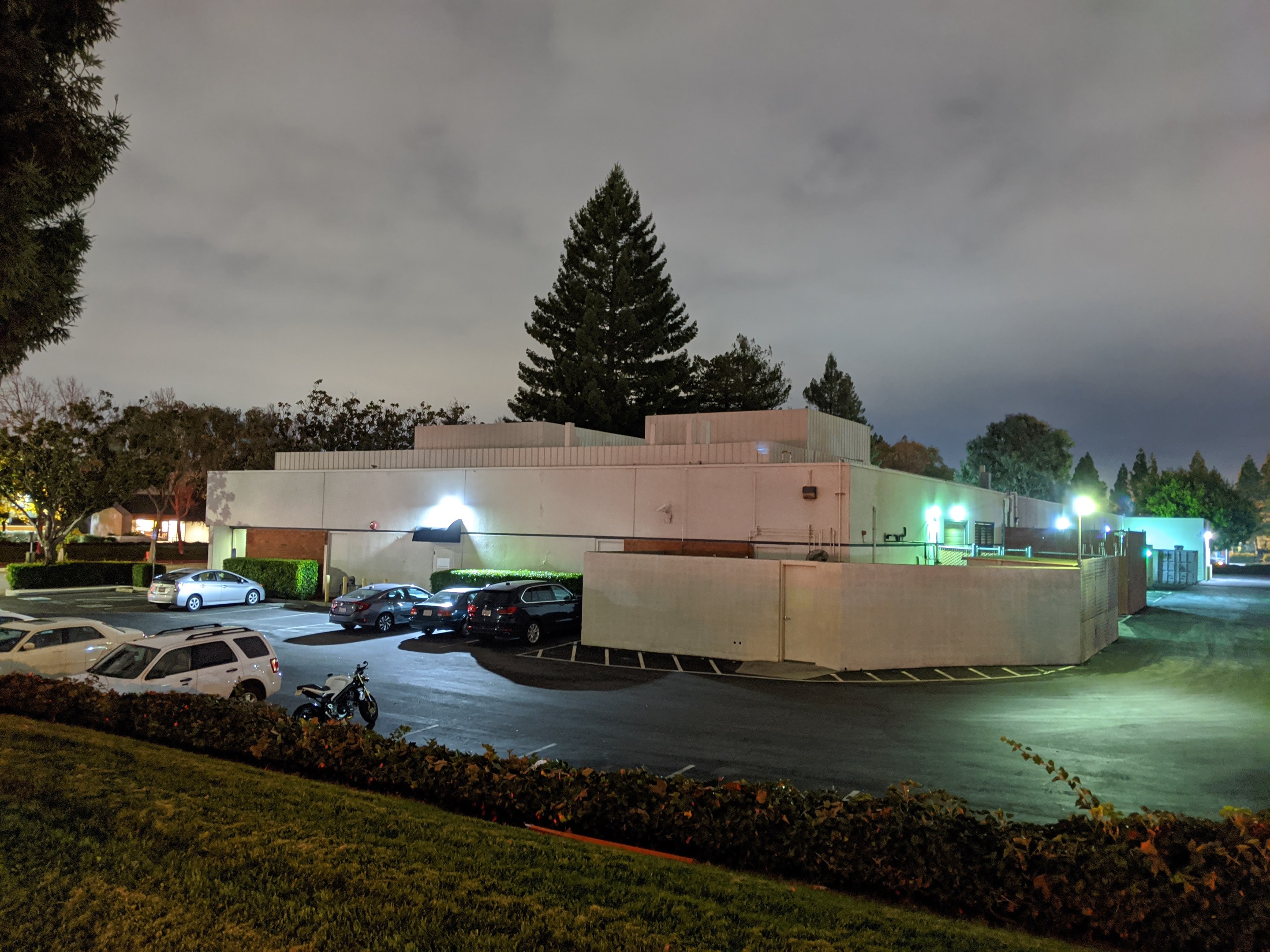 Parkplatz bei Nacht mit geparkten Autos, Gebäuden, Straßenlaternen, fahrenden Autos, Grünflächen und bewölktem Himmel.