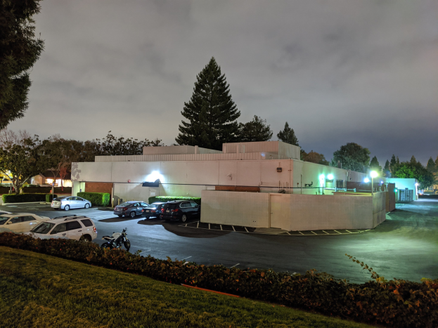 Parkplatz bei Nacht mit geparkten Autos, Gebäuden, Straßenlaternen, fahrenden Autos, Grünflächen und bewölktem Himmel.