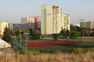 Eine Gruppe von Menschen, die Tennis auf einem Court spielen, umgeben von hohen Gebäuden, üppiger Vegetation, Laternenmasten und Fahrzeugen auf einer nahen Straße, mit einem sichtbaren Himmel darüber.
