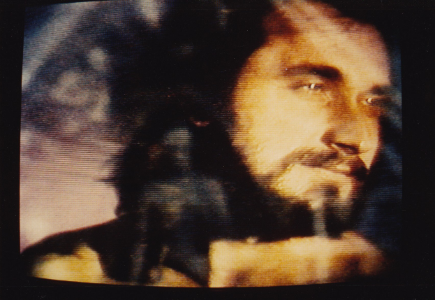 Ein Mann mit Bart und Schnurrbart auf einem TV-Bildschirm, dunkler Hintergrund, ernster Gesichtsausdruck, weißes Hemd, direkter Blick.