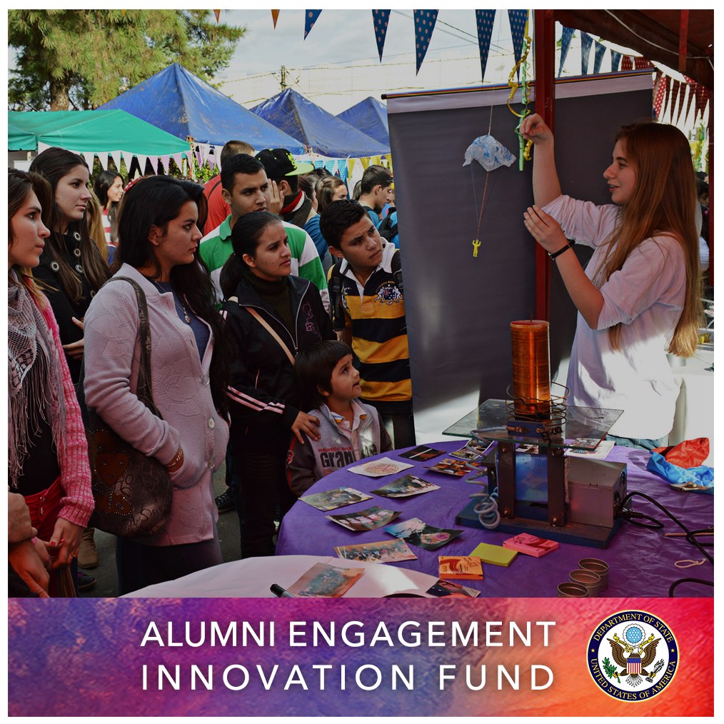 Eine Gruppe von Menschen steht um einen Tisch mit einer "Alumni Engagement Innovation Fund"-Aufschrift, umgeben von Zelten, Bäumen und einem klaren blauen Himmel, mit Text und einem Logo unten.