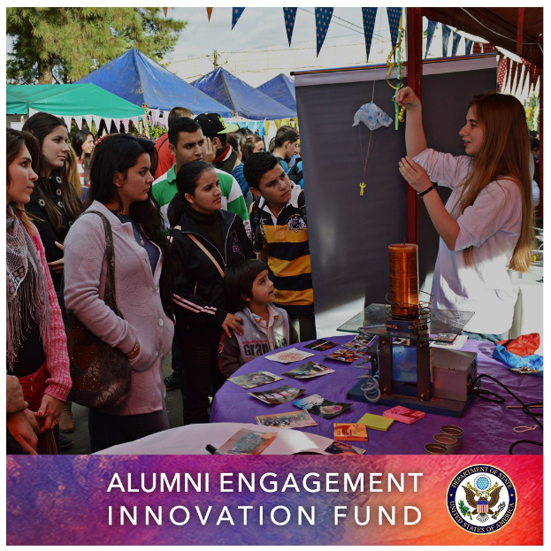 Eine Gruppe von Menschen steht um einen Tisch mit einer "Alumni Engagement Innovation Fund"-Aufschrift, umgeben von Zelten, Bäumen und einem klaren blauen Himmel, mit Text und einem Logo unten.