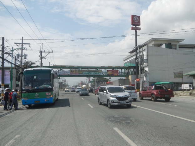 Ein Stadtbus fährt auf einer Straße mit hohen Gebäuden, Strommasten und einer Gruppe von Menschen auf dem Gehweg, mit einem Bogen, Bäumen und einem bewölkten Himmel im Hintergrund.