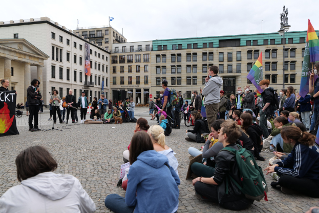 Eine Gruppe von Menschen, die auf dem Boden sitzen, vor einer Menge mit Fahnen und Spruchbändern bei einer Anti-Schwulen-Demo in Berlin. Im Hintergrund sind eine Statue, Gebäude und ein Mikrofonständer zu sehen.