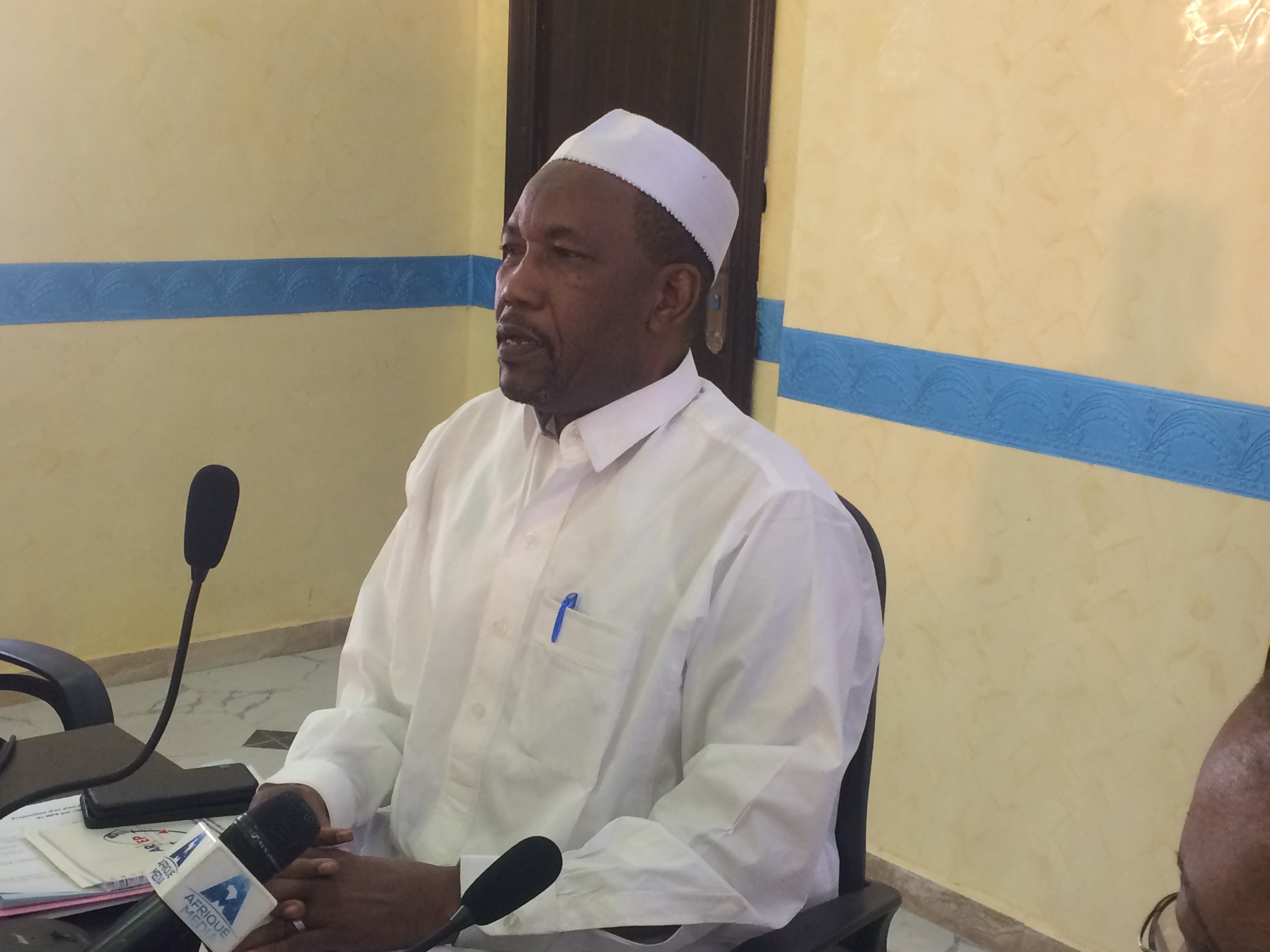 Ein Mann in einem weißen Kleid und Hut sitzt an einem Tisch mit Mikrofonen, spricht, während Papiere und Gegenstände auf dem Tisch sichtbar sind, mit einer anderen Person mit Brille rechts von ihm, in einem Raum mit einer Wand und einer Tür im Hintergrund.