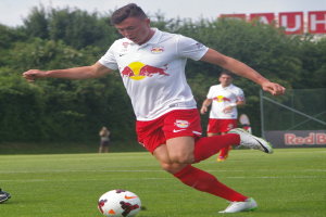 Ein Mann in einem Fussballtrikot kickt einen Ball auf einem grünen Feld mit Bäumen und einem klaren blauen Himmel im Hintergrund, mit einem Schild mit der Aufschrift "RB Leipzig" im Bild.