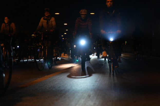 Eine Gruppe von Menschen, die nachts auf Fahrrädern durch eine Stadtstraße fahren und Helme tragen, beleuchtet von Stadtlichtern.