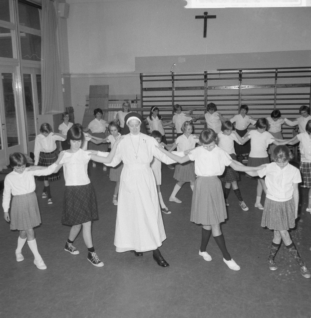 Schwarzes und weißes Bild von Kindern in Schuluniformen, die in einem Raum mit einer wandmontierten Kreuzt tanzen, Glastüren mit Vorhängen und Deckenleuchten.