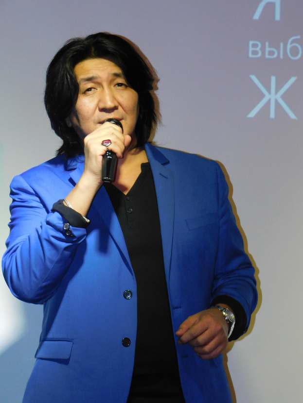 Ein Mann in einem blauen Anzug und schwarzer Krawatte hält ein Mikrofon und steht vor einem Bildschirm mit Text, trägt ein weißes Hemd und hat einen ernsten Gesichtsausdruck.