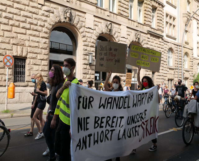 Gruppe von Menschen bei einer Anti-Kriegs-Demonstration in Berlin, die eine Straße mit Schildern, Bannern und Fahrrädern entlanggeht, an einem Gebäude mit Fenstern, Bögen, Säulen und Skulpturen sowie Bäumen im Hintergrund vorbeikommt.