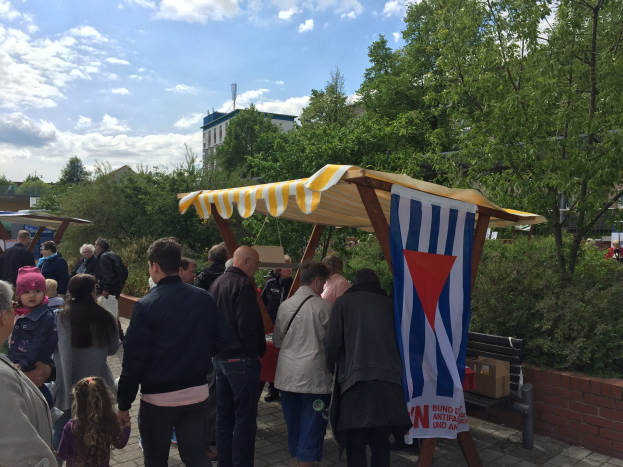 Gruppe von Menschen versammelt sich vor einem Zelt mit einer Fahne, in der Nähe einer Bank und einer Tafel mit Text, vor Bäumen, Gebäuden und einem bewölkten Himmel.