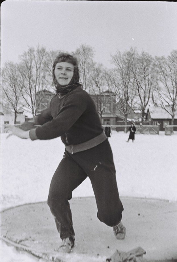 Schwarz-weiß-Foto einer Frau, die auf einem Skateboard im Schnee fährt, mit Menschen, Bäumen, Gebäuden und einem klaren Himmel im Hintergrund, mit Text unten.