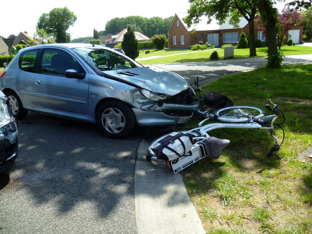 Ein Auto ist auf der Straße gegen einen Zaun geprallt, daneben liegt ein Fahrrad auf dem Rasen, Häuser und Bäume im Hintergrund vor einem klaren blauen Himmel.