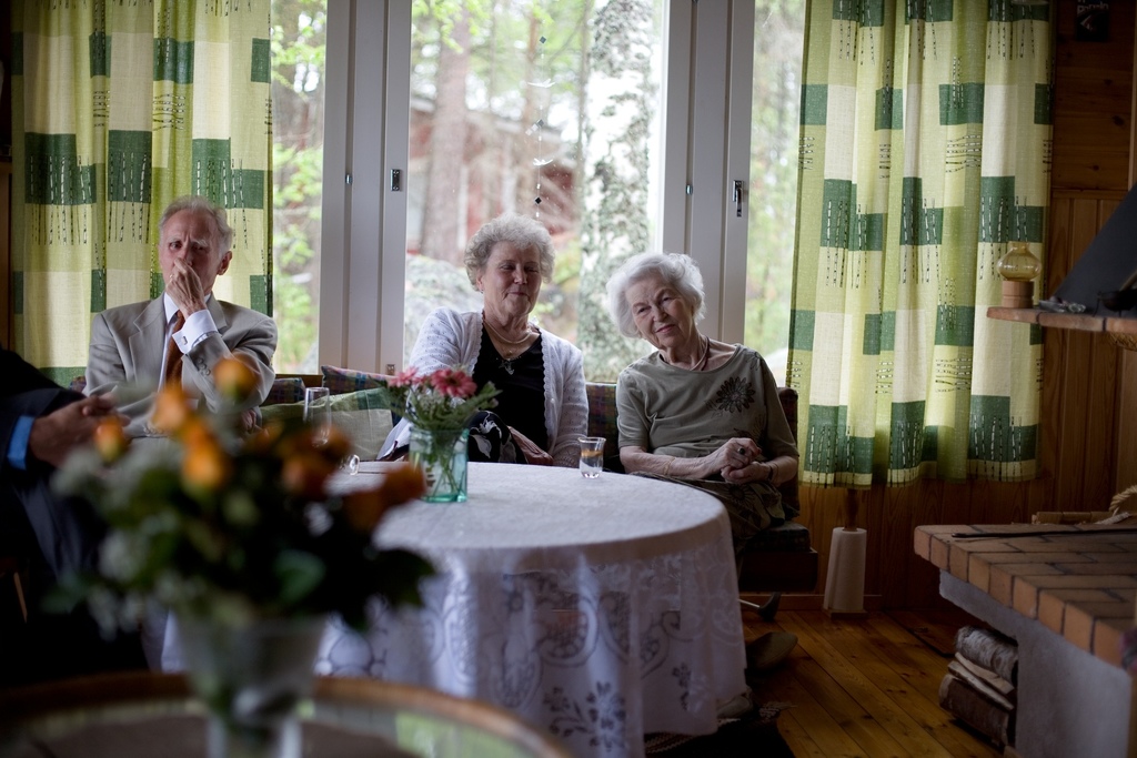 Eine Gruppe älterer Menschen sitzt auf einem Sofa mit einem Tisch und einer Blumenvase davor, eine Glastür und Vorhänge im Hintergrund.