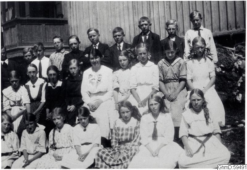 Schwarzes und weißes Foto einer Gruppe von Kindern, die vor einem Gebäude mit Pflanzen im Hintergrund posieren, mit der Beschriftung "1910-1911 Schulkinder" unten.