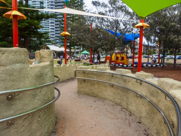 Spielplatz mit einem grün-gelben Dach, umgeben von Geländern, Pfählen, Zelten, Bäumen, Fahrzeugen, Menschen und Gebäuden unter einem klaren blauen Himmel.