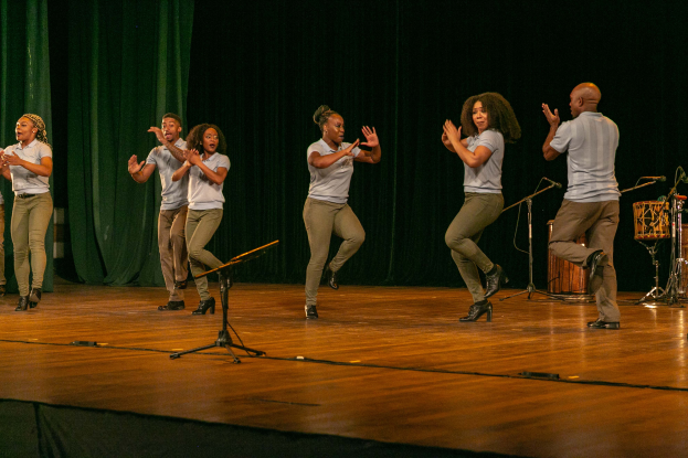 Eine Gruppe von Menschen auf einer Bühne mit Mikrofonen und Musikinstrumenten, die klatschen und feiern, mit einem Vorhang im Hintergrund.