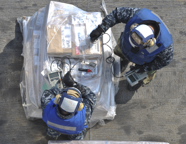 Zwei Männer in blauen Uniformen und Helmen arbeiten an einem großen Drohnenmodell und halten verschiedene Gegenstände, mit einer Plastikabdeckung auf dem Boden.