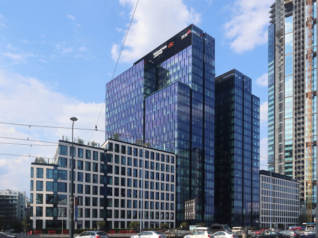 Großes Gebäude mit vielen Fenstern, identifiziert als Deutsche Bank Hauptsitz in Frankfurt, Deutschland, umgeben von städtischen Elementen einschließlich Straßenlaternen, Fahrzeugen, Bäumen und einem bewölkten Himmel.