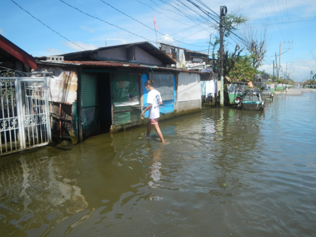 Ein Mann in einem weißen T-Shirt und Shorts watet durch eine überflutete Straße in Manila, umgeben von Häusern, Bäumen, Polen, Drähten und Fahrzeugen unter einem sichtbaren Himmel.