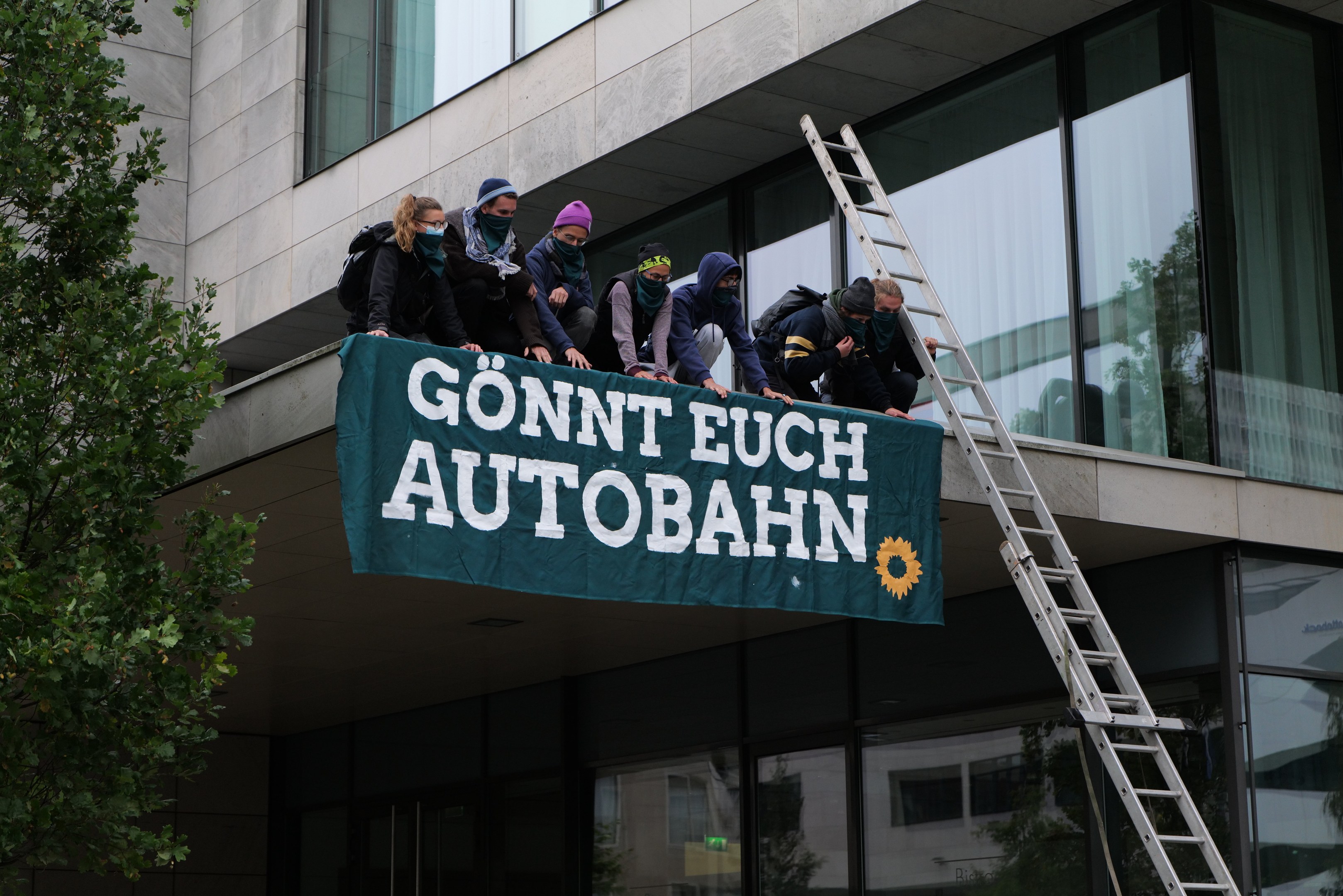 Menschen auf einem Gebäude mit einem Banner, mit einem Baum links und Glasfenstern im Hintergrund, wahrscheinlich protestieren sie für die deutsche Regierung.