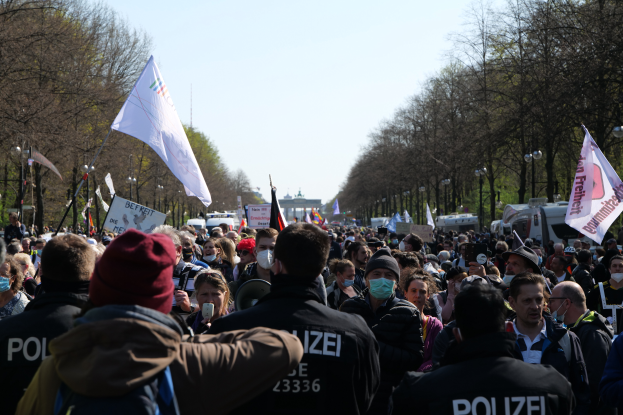 Eine große Gruppe von Menschen steht vor einer Menge von Polizisten, einige tragen Mützen und Masken, während sie auf einer Demonstration in Berlin, Deutschland, mit Schildern, Fahnen, Laternenpfählen, Bäumen, Fahrzeugen, einem Gebäude und dem Himmel im Hintergrund demonstrieren