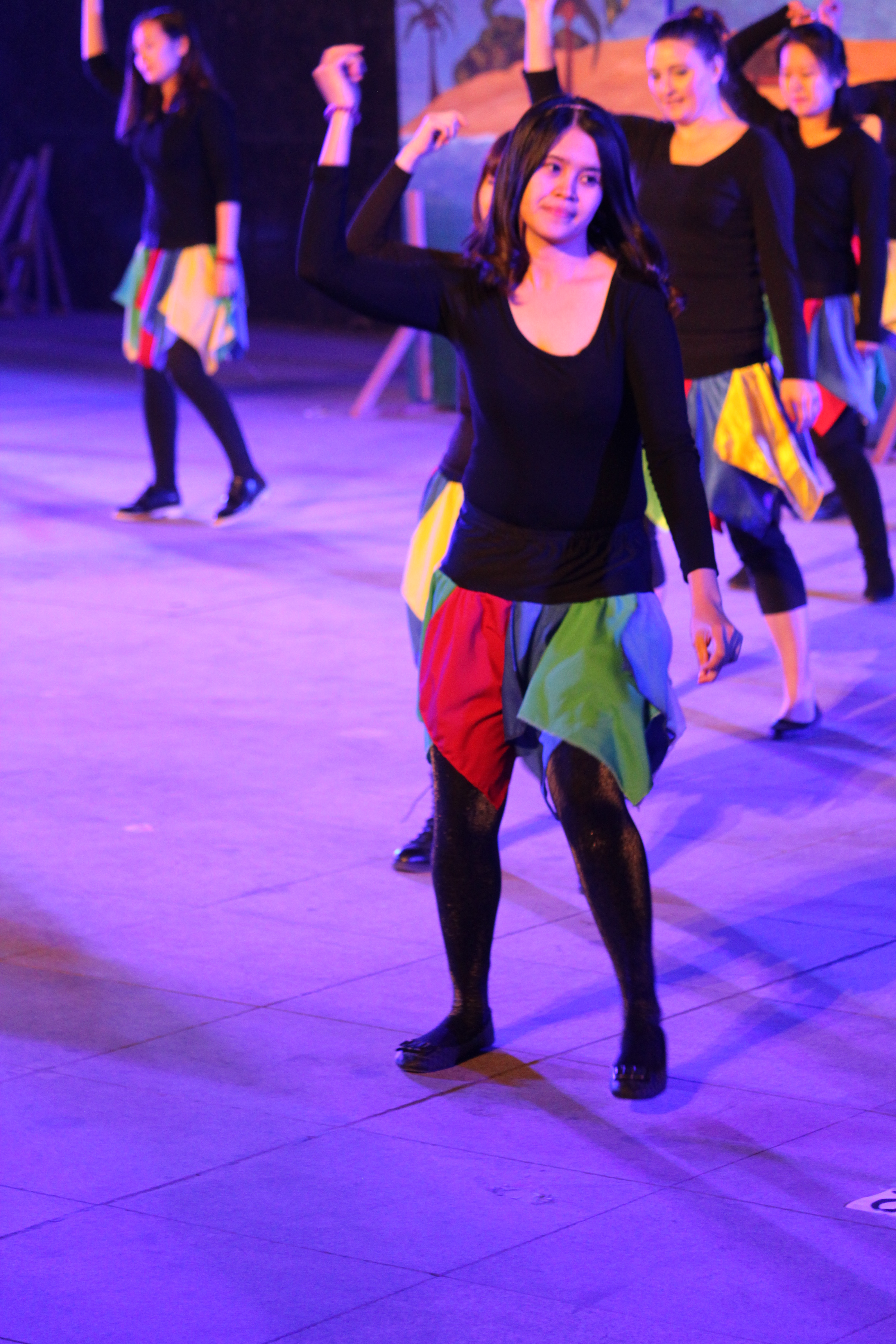 Eine Gruppe junger Frauen tanzt auf einer Bühne, mit einem Gemälde im Hintergrund, alle tragen Schuhe.