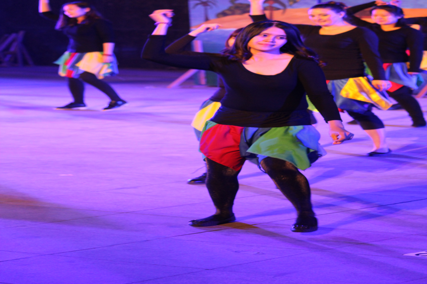 Eine Gruppe junger Frauen tanzt auf einer Bühne, mit einem Gemälde im Hintergrund, alle tragen Schuhe.