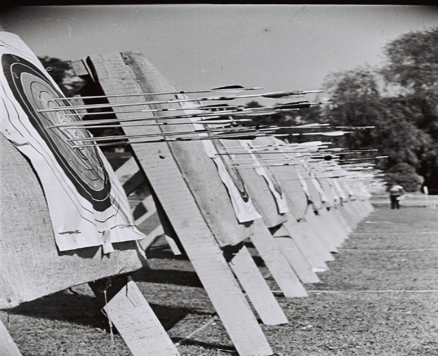 Schwarzes und weisses Foto eines Bogenschiessziels mit Pfeilen in der Mitte, umgeben von Bäumen und einer Person im Hintergrund, mit sichtbarem Boden unten.