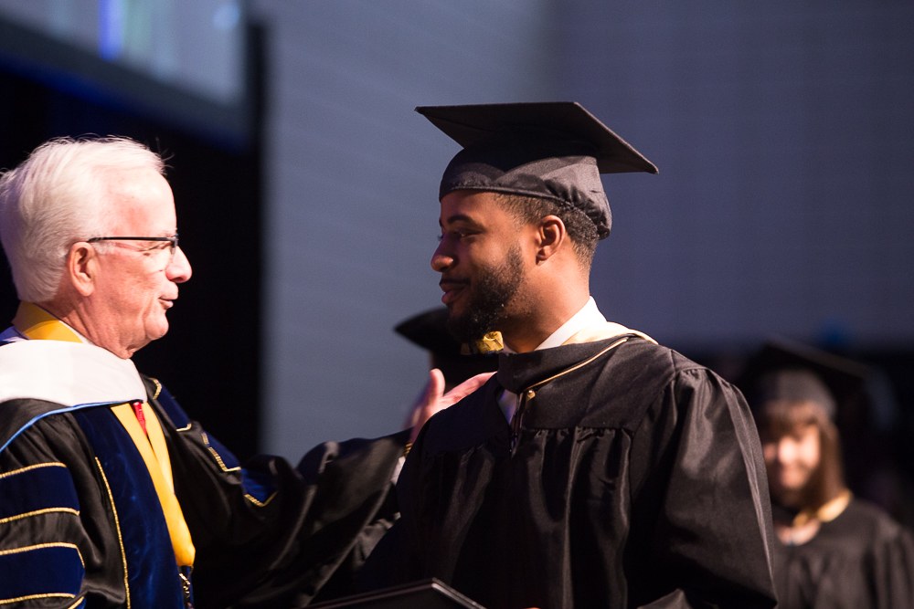 Zwei Männer, einer in einer Graduationskapelle und -robe mit einem Buch in der Hand, schütteln sich die Hand und lächeln, während andere im Hintergrund mit einem Bildschirm zu sehen sind.